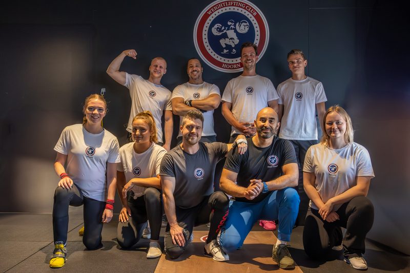 Elf leden van Weightlifting Club Hoorn in clubshirt, samen op de foto voor het clublogo op de muur van de zaal.