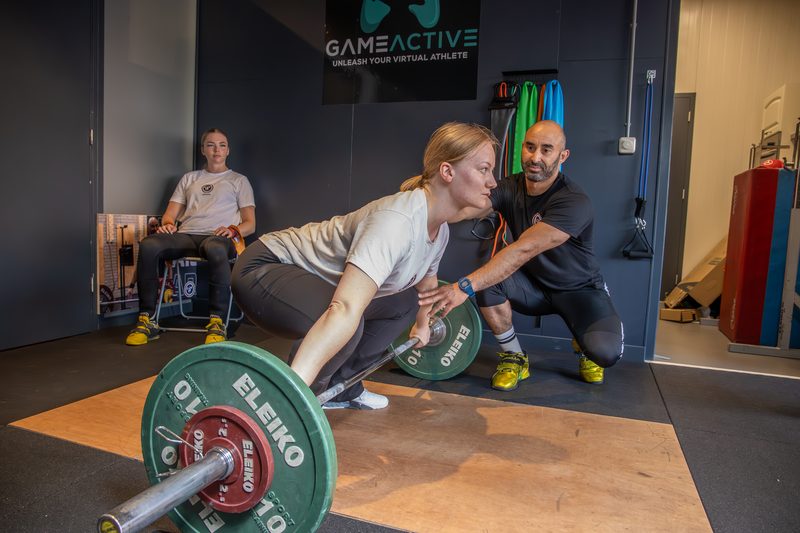 Omar Khathiri hurkt naast een lifter en wijst haar de startpositie aan boven de stang.