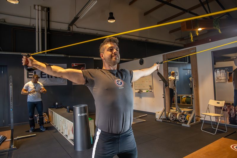 Marco houdt een elastiek gestrekt boven zijn hoofd tijdens een mobiliteitsoefening in de zaal.