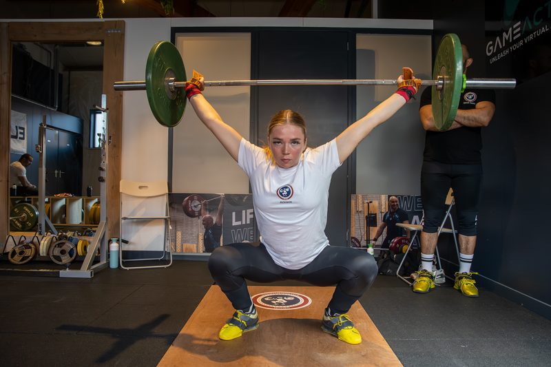 Een lifter komt omhoog uit de hurkzit met de stang boven het hoofd, tijdens een training in de zaal.
