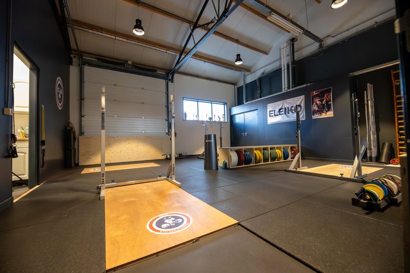 De zaal van Weightlifting Club Hoorn met open roldeur, heftplatform en gekleurde platen langs de wand.